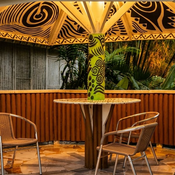Outdoor seating area with a patterned umbrella, round table, and wicker chairs, surrounded by lush greenery.