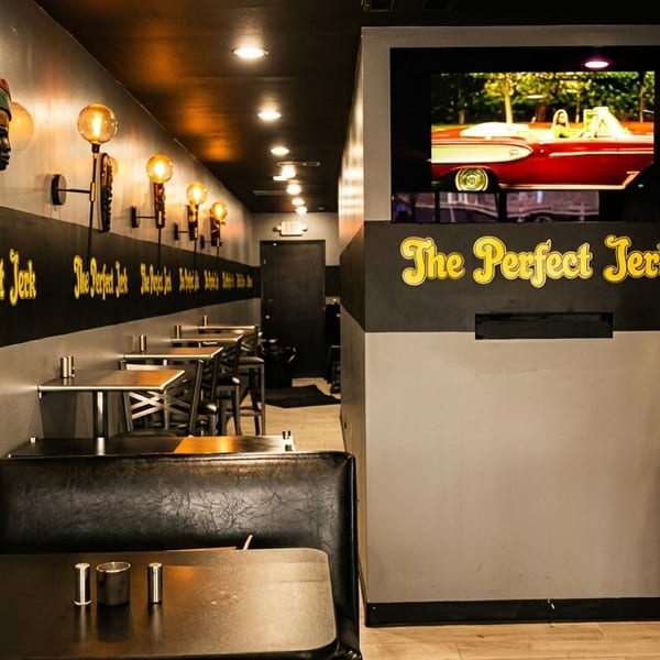 Interior of The Perfect Jerk restaurant with booths, tables, wall sconces, and a TV screen.